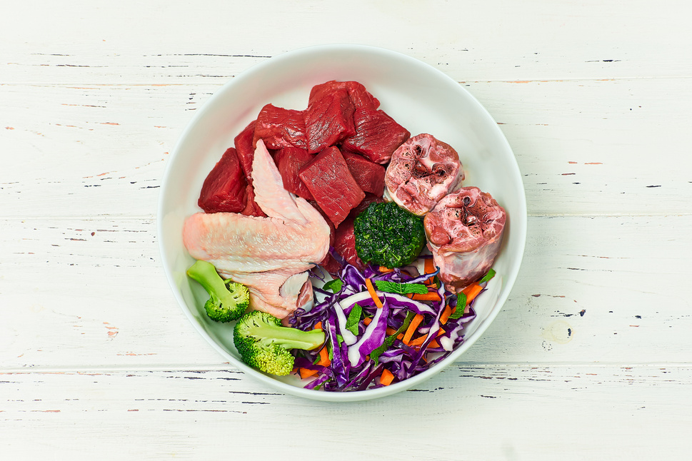 Pet food. Bowl of natural raw dog food on white wooden background. Beef meat, turkey neck, chicken wing and vegetables. BARF diet for dog.